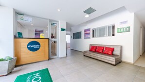 Un espace d’accueil d’hôtel comprenant un comptoir de réception, un canapé avec des coussins rouges et un tapis vert orné du nom de l’hôtel.