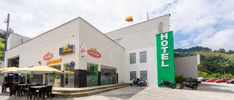 Un hotel con un llamativo letrero verde que dice 'HOTEL', zona de estar al aire libre con sombrillas y un aparcamiento con motocicletas.