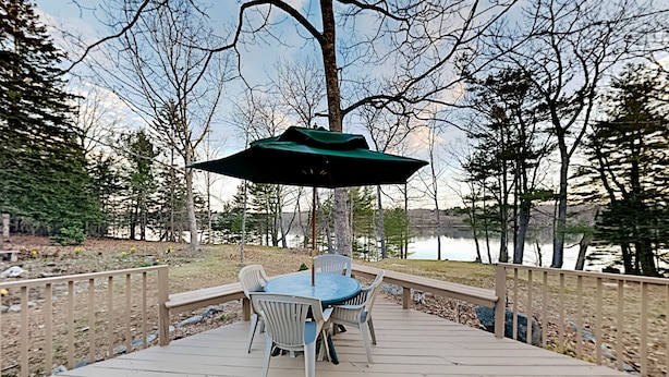 Eine Holzterrasse mit Tisch und Stühlen, umgeben von Bäumen und einem Gewässer.
