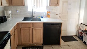 A compact kitchen with wooden cabinets, a dishwasher, a coffee maker, and a window with blinds. - Monthly winter rates. Ground floor. Heart of Surf City. 515 N. Shore Dr. Apt. #3 (Surf City)