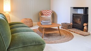 A cozy living room with a green sofa, a wicker chair, a small round table, a lamp, and a wood-burning stove.
