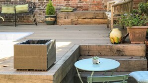 A patio with a metal chair, a small table, a planter with a plant, and a brick wall in the background.