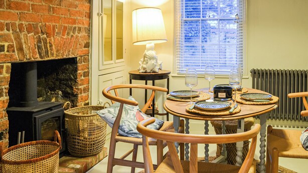 A cozy room with a brick fireplace, wicker baskets, a table with plates and glasses, and a window with blinds.