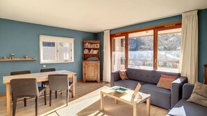 A living room with a dining table, chairs, a sofa, a bookshelf, and a window with curtains. - APARTMENT WITH POOL ACCESS - ST JEAN D'AULPS RESORT - 6 Pers / 60m² - Riam D2 (Saint-Jean-d´Aulps)