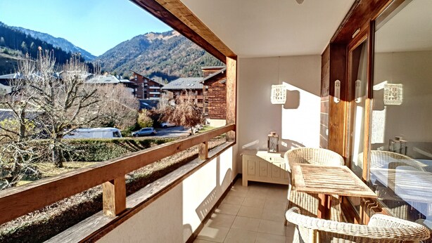 A balcony with a view of a mountain and a village. There is a wicker chair and a small table with a lantern. - APARTMENT WITH POOL ACCESS - ST JEAN D'AULPS RESORT - 6 Pers / 60m² - Riam D2 (Saint-Jean-d´Aulps)