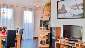 A room with a dining table, chairs, a TV, a bookshelf, and a painting of a lighthouse. - Vacation home Bodden - semi-detached house (Saal)