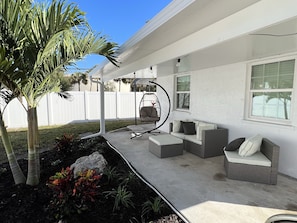 Relax in a serene outdoor space with a sofa, chair, and swing chair, surrounded by lush plants and a palm tree.