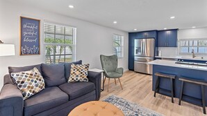 Ein modernes Wohnzimmer mit einer Couch, einem gemusterten Kissen, einem grauen Sessel und einer Küchenzeile mit einem Edelstahlkühlschrank und blauen Schränken.