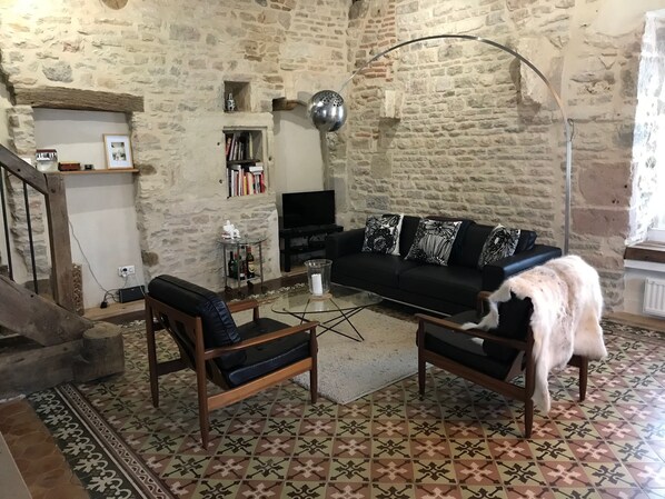 Ein gemütliches Wohnzimmer mit Steinwand, einer schwarzen Couch mit gemusterten Kissen, einem Holztisch und einem schwarzen Ledersessel.