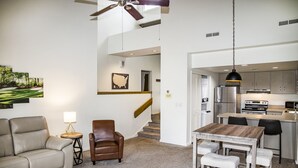Un vaste espace de vie comprenant un canapé, une table basse et une table à manger avec des bancs. Il y a également une cuisine équipée d’appareils électroménagers modernes et un escalier menant à l’étage supérieur.