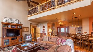A spacious living room with a large television, a fireplace, a dining area with wooden furniture, and a staircase with a wooden railing. - Mossy Path Hideaway - Blowing Rock area home with easy access and ping pong! (Blowing Rock)