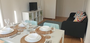 A dining area with a white table set for four, surrounded by a black sofa with colorful pillows, a white entertainment unit with a TV, and a small plant.