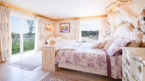 A spacious bedroom with a large bed, a nightstand, and a dresser. There is a window with curtains, a framed picture on the wall, and a view of the outdoors.
