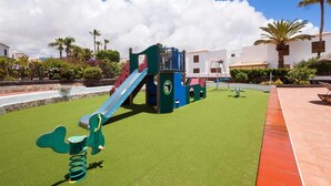 Un'area giochi con scivolo e strutture di arrampicata su erba sintetica, circondata da palme e edifici.