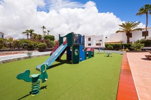 Une aire de jeux avec un toboggan et des structures d’escalade sur une pelouse artificielle, entourée de palmiers et d’immeubles.