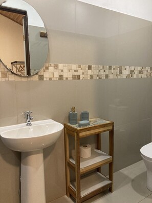 Un baño con lavamanos blanco, espejo, un pequeño estante de madera con artículos de aseo y salpicadero de azulejos.
