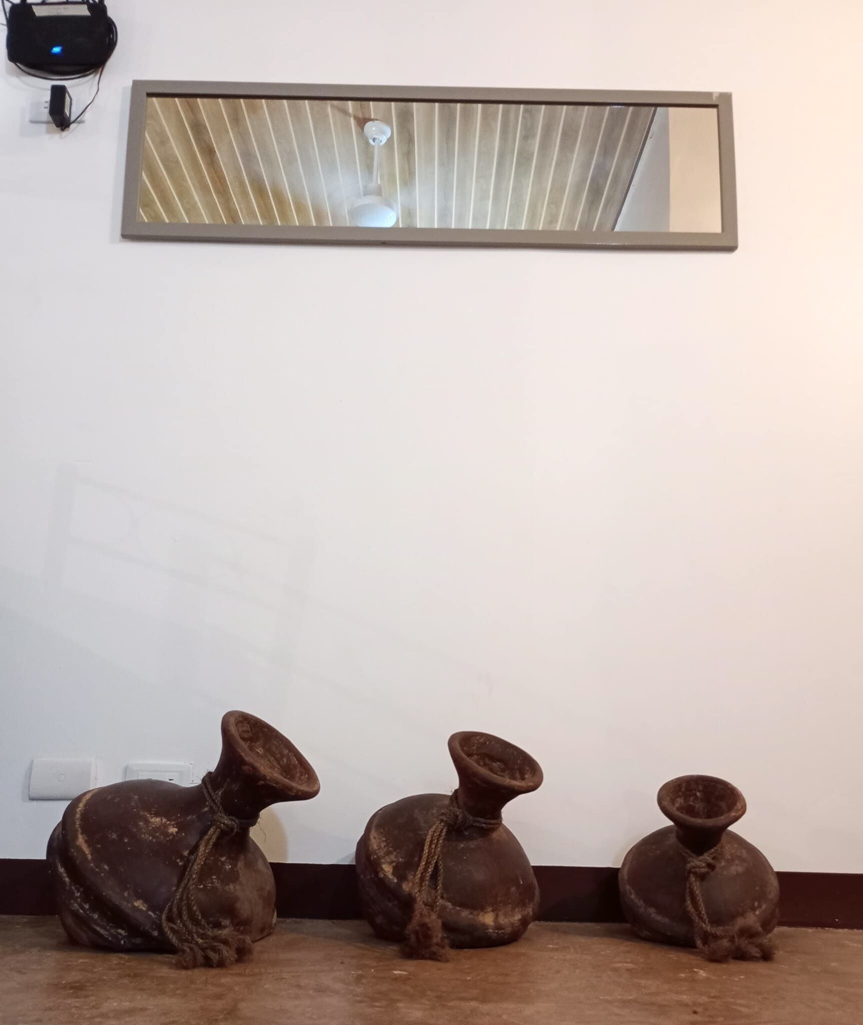 Tres piezas de cerámica sobre el suelo frente a una pared blanca, con un ventilador de techo y un espejo arriba.