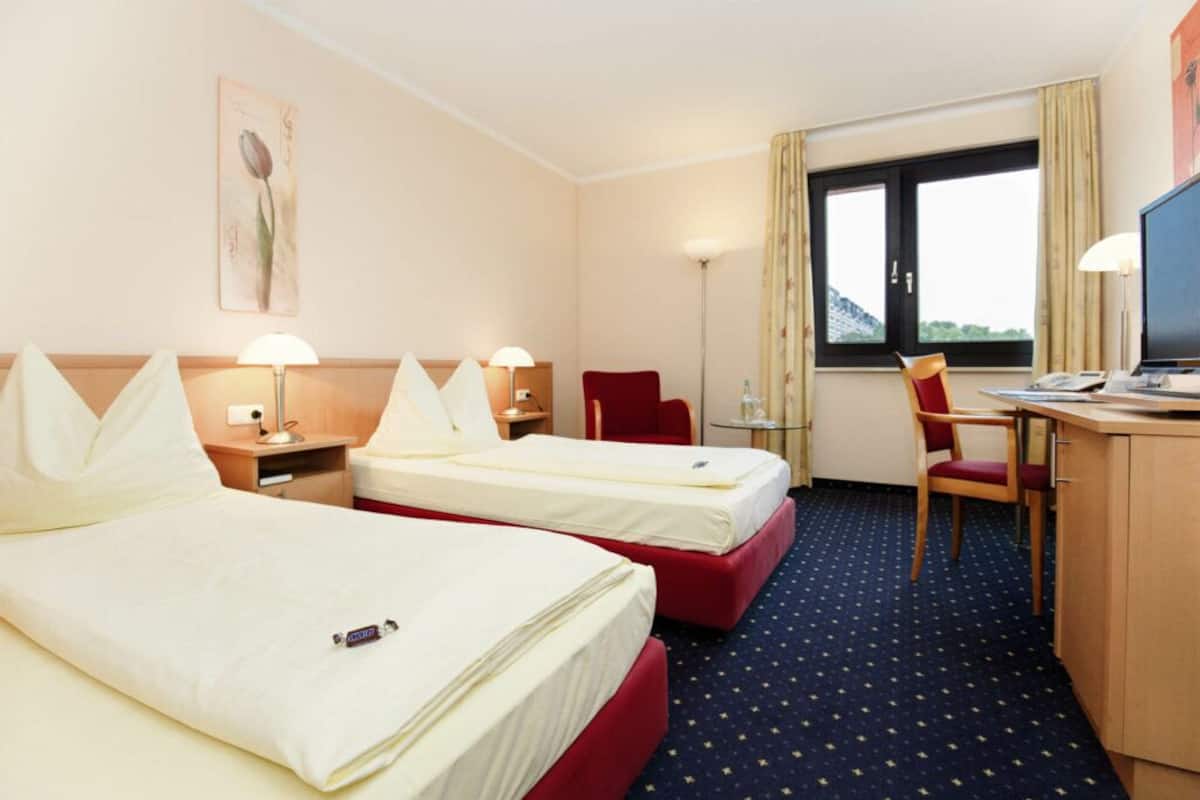 Ein Hotelzimmer mit zwei Betten, einem Schreibtisch, einem Stuhl, einem Fernseher und einem Fenster mit Vorhängen.
