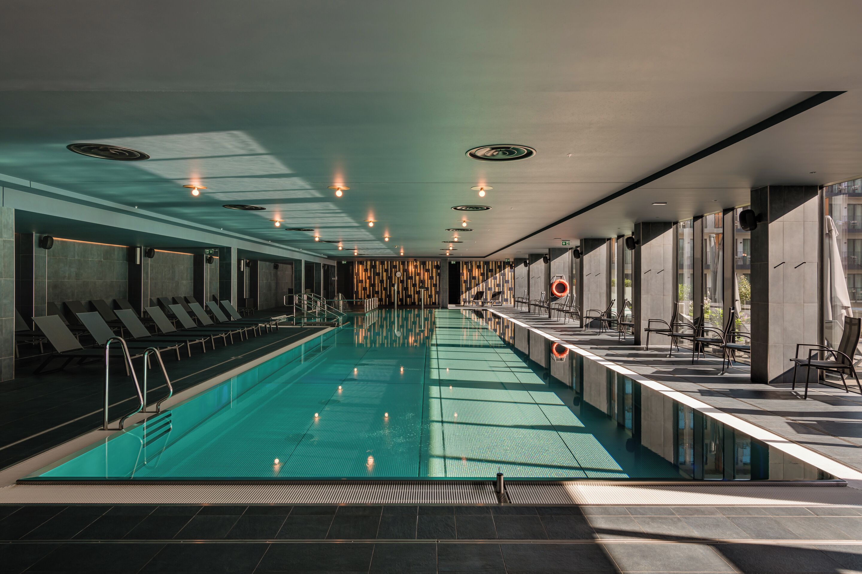 A modern indoor swimming pool with a long, narrow design, surrounded by lounge chairs and a seating area with tables.