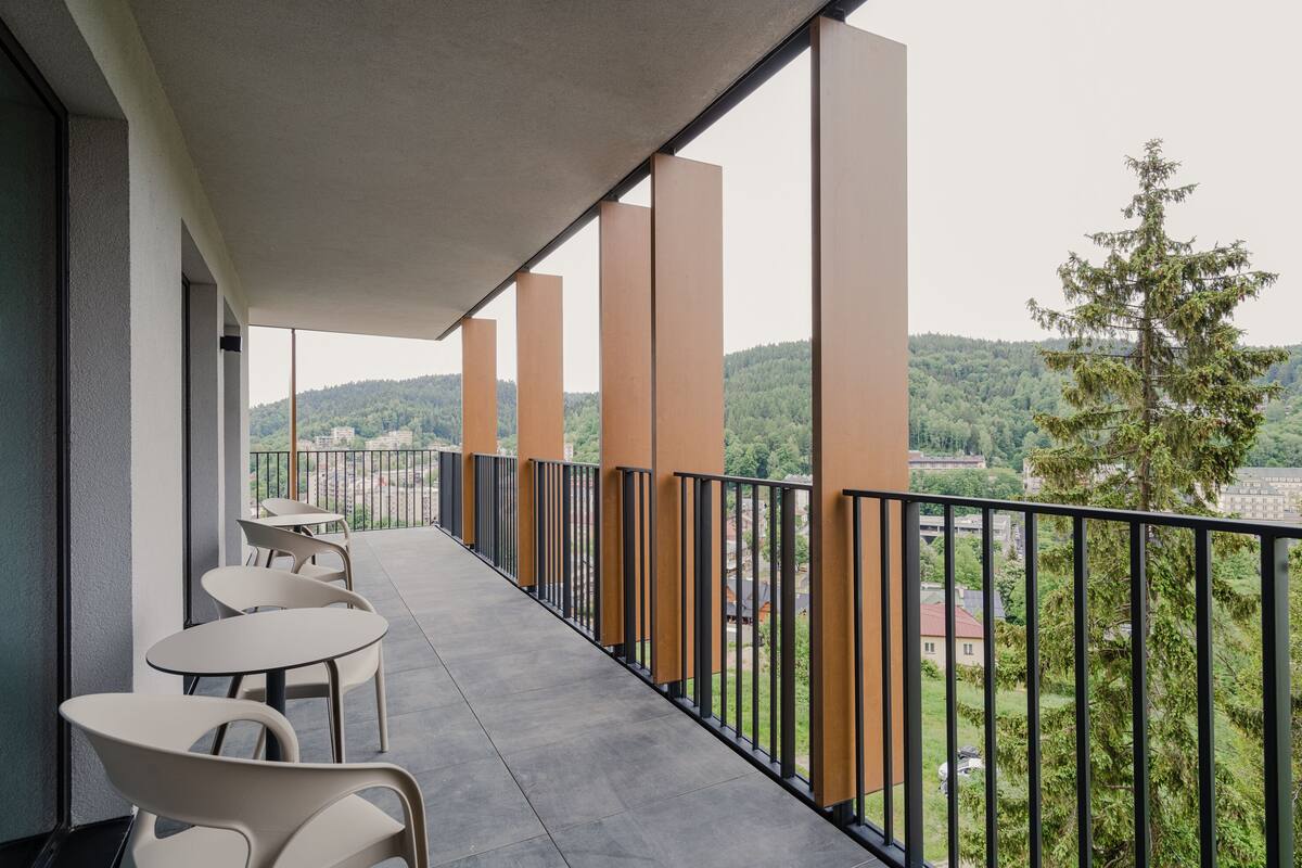 Un balcon agrémenté de tables et de chaises, d’où l’on peut admirer une zone boisée et des bâtiments.