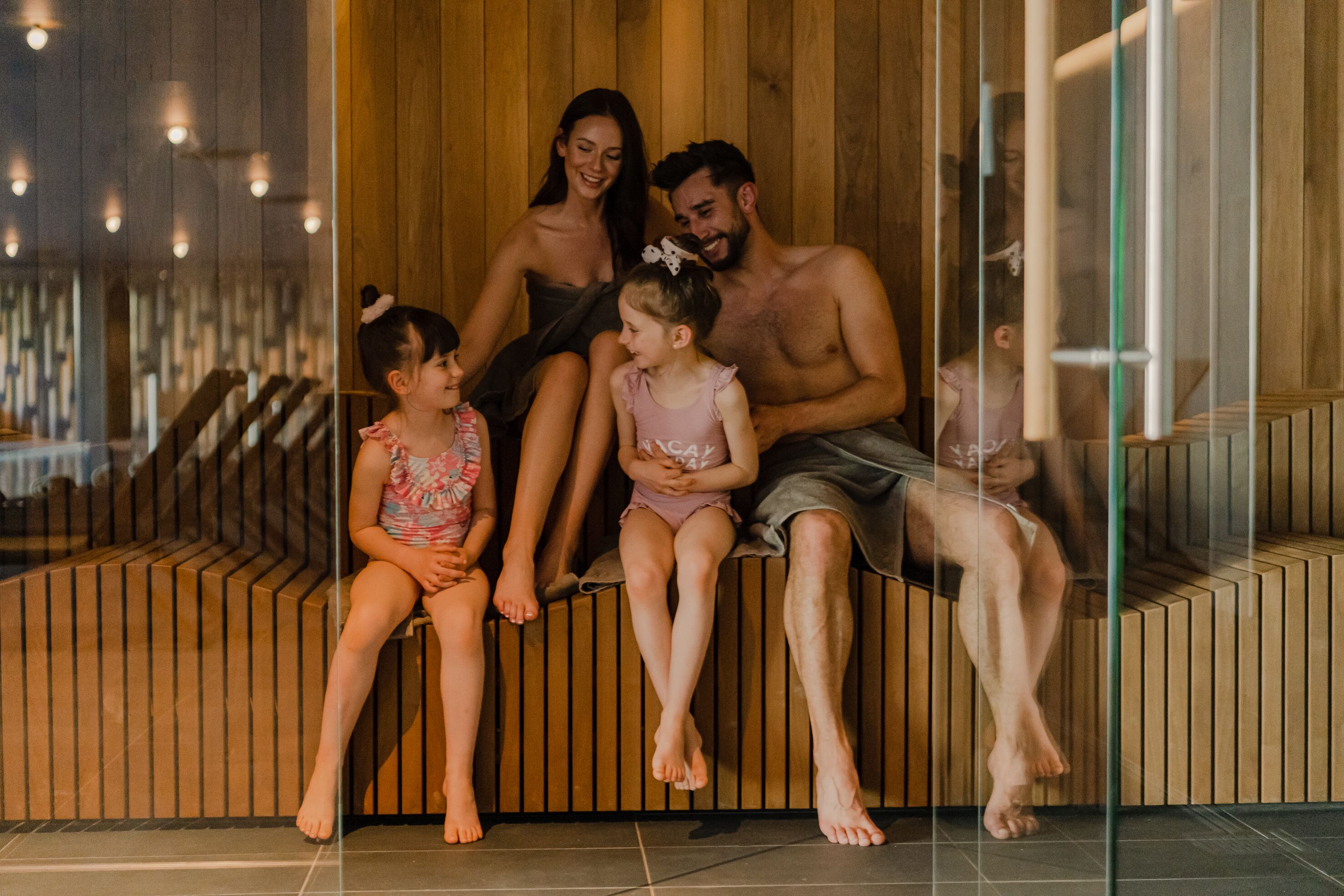A family of four seated on a wooden bench in a modern, well-lit indoor space with a wooden wall and glass partition.