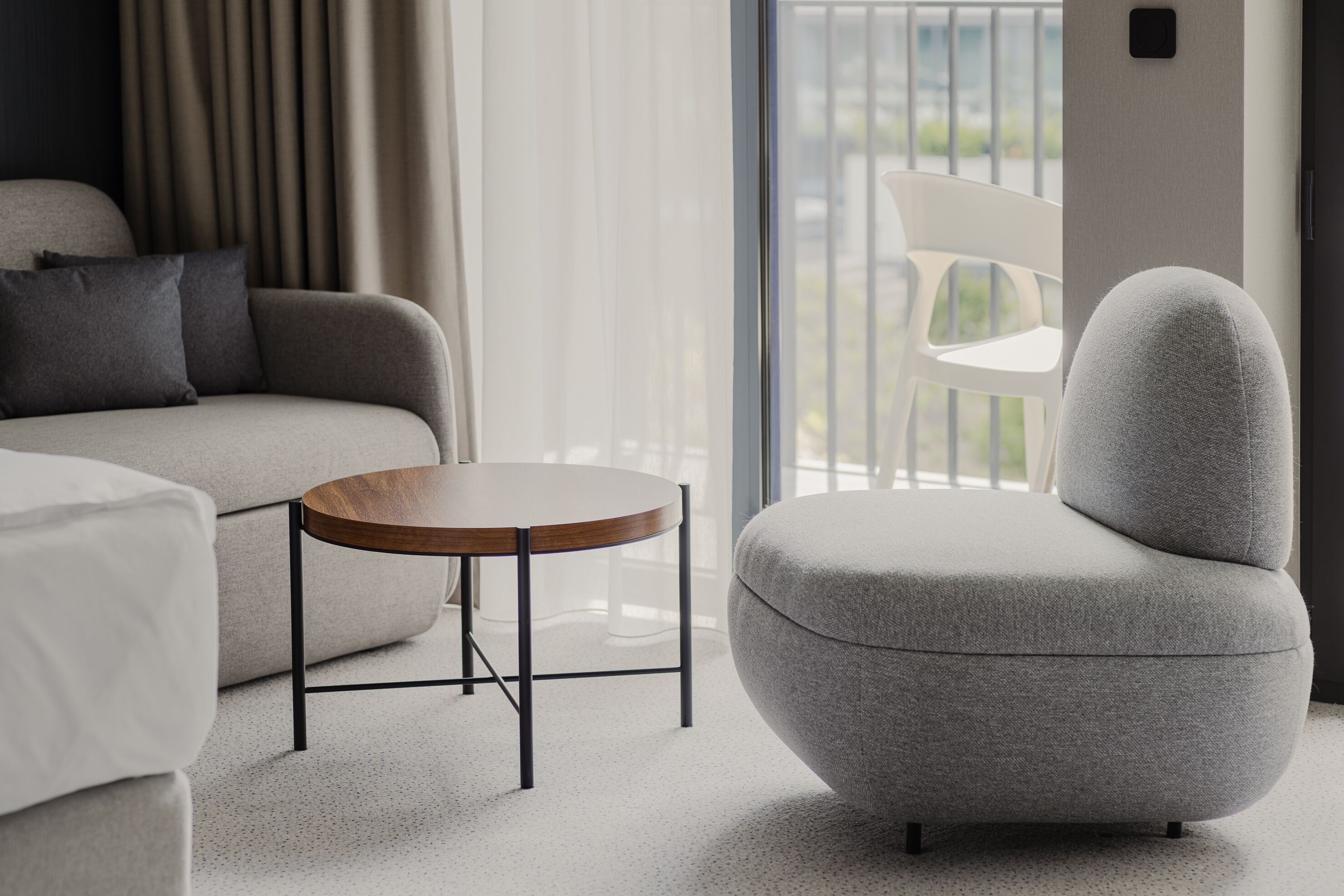 A modern hotel room with a grey sofa, a round wooden table, and a grey armchair.