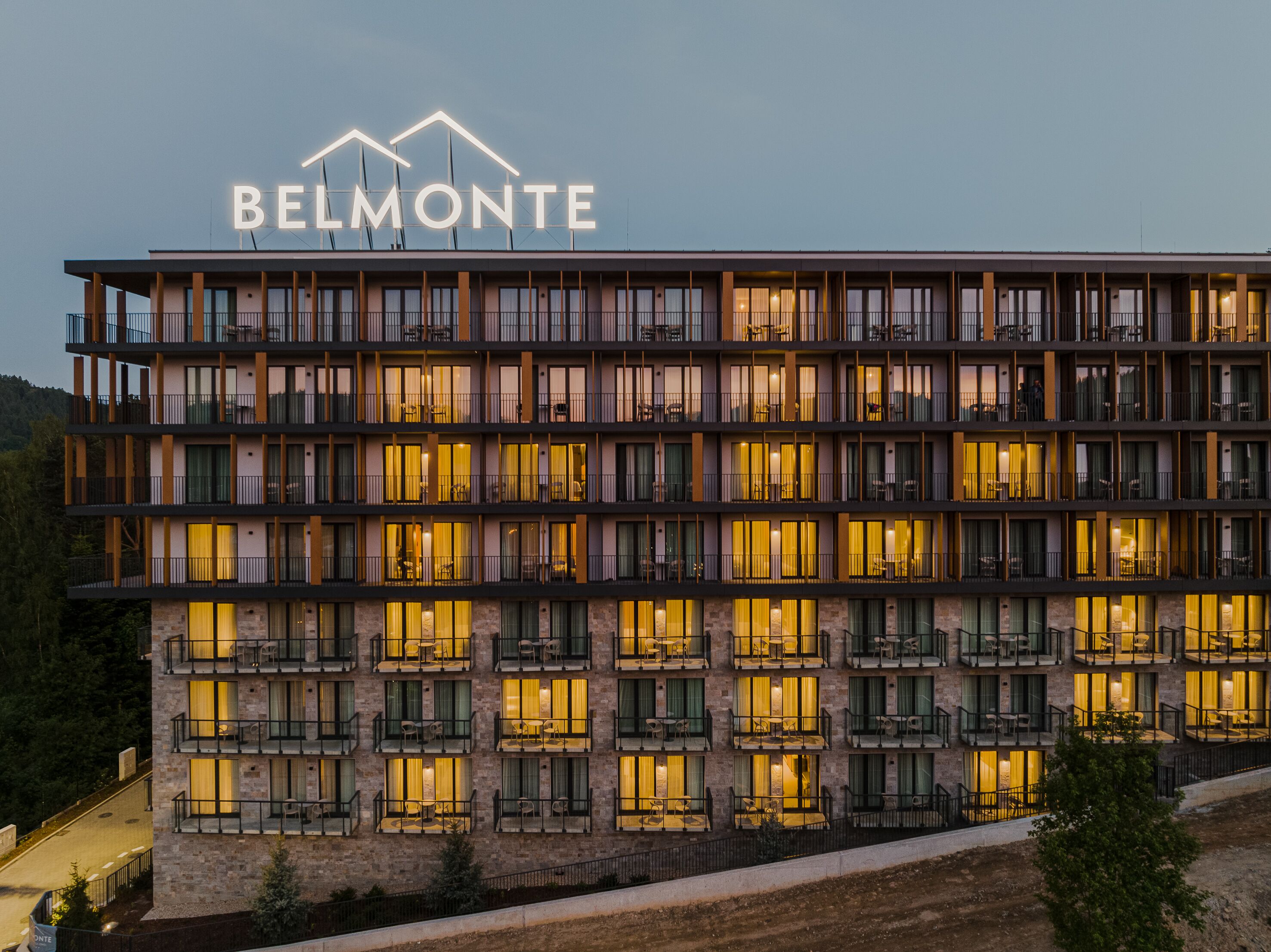 Un hôtel à plusieurs étages, reconnaissable à son grand panneau « BELMONTE », doté de balcons avec des espaces salons extérieurs et d’une façade en pierre.