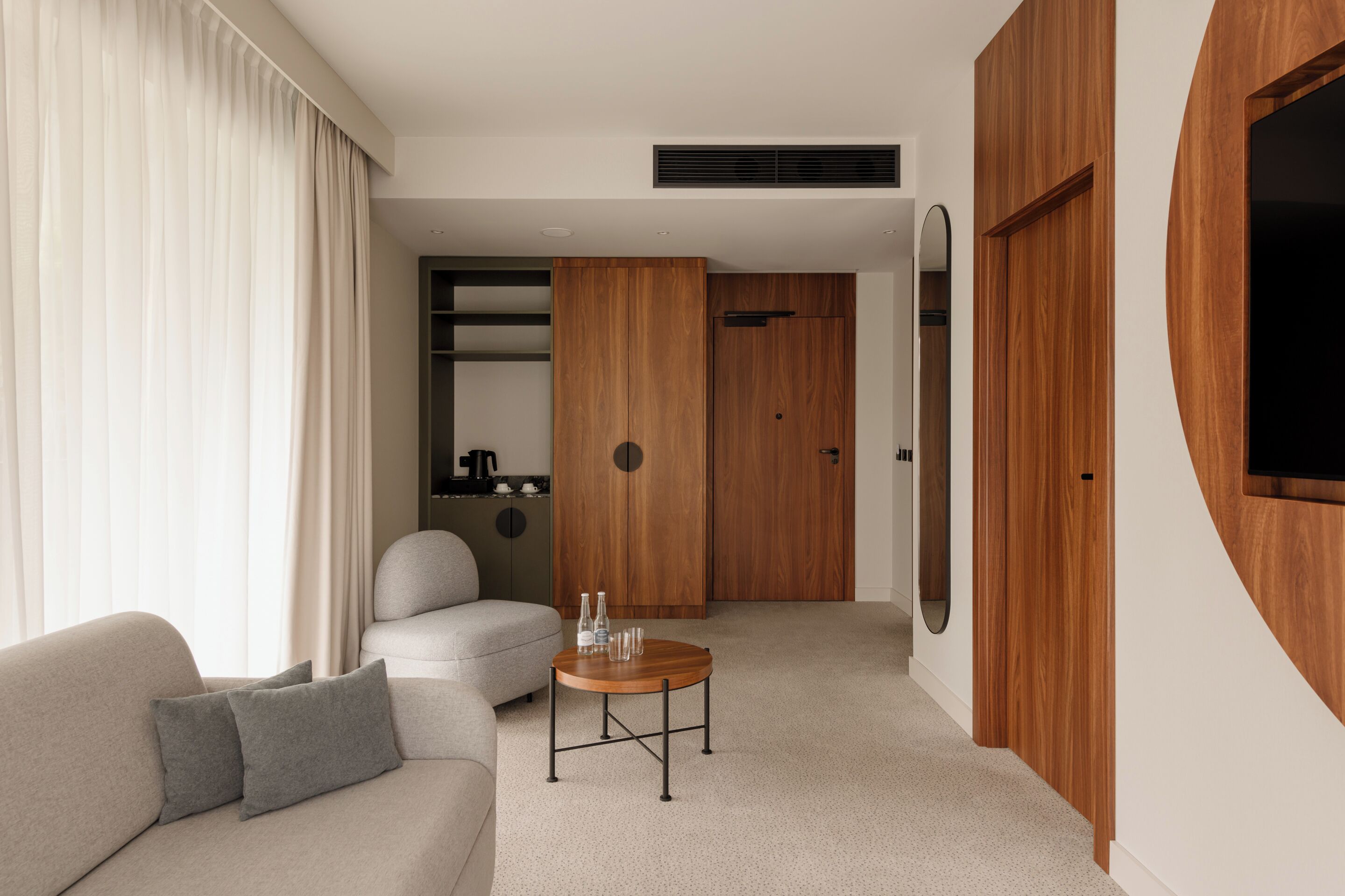 A modern hotel room with a sofa, a round wooden coffee table, a wardrobe, and a television mounted on the wall.