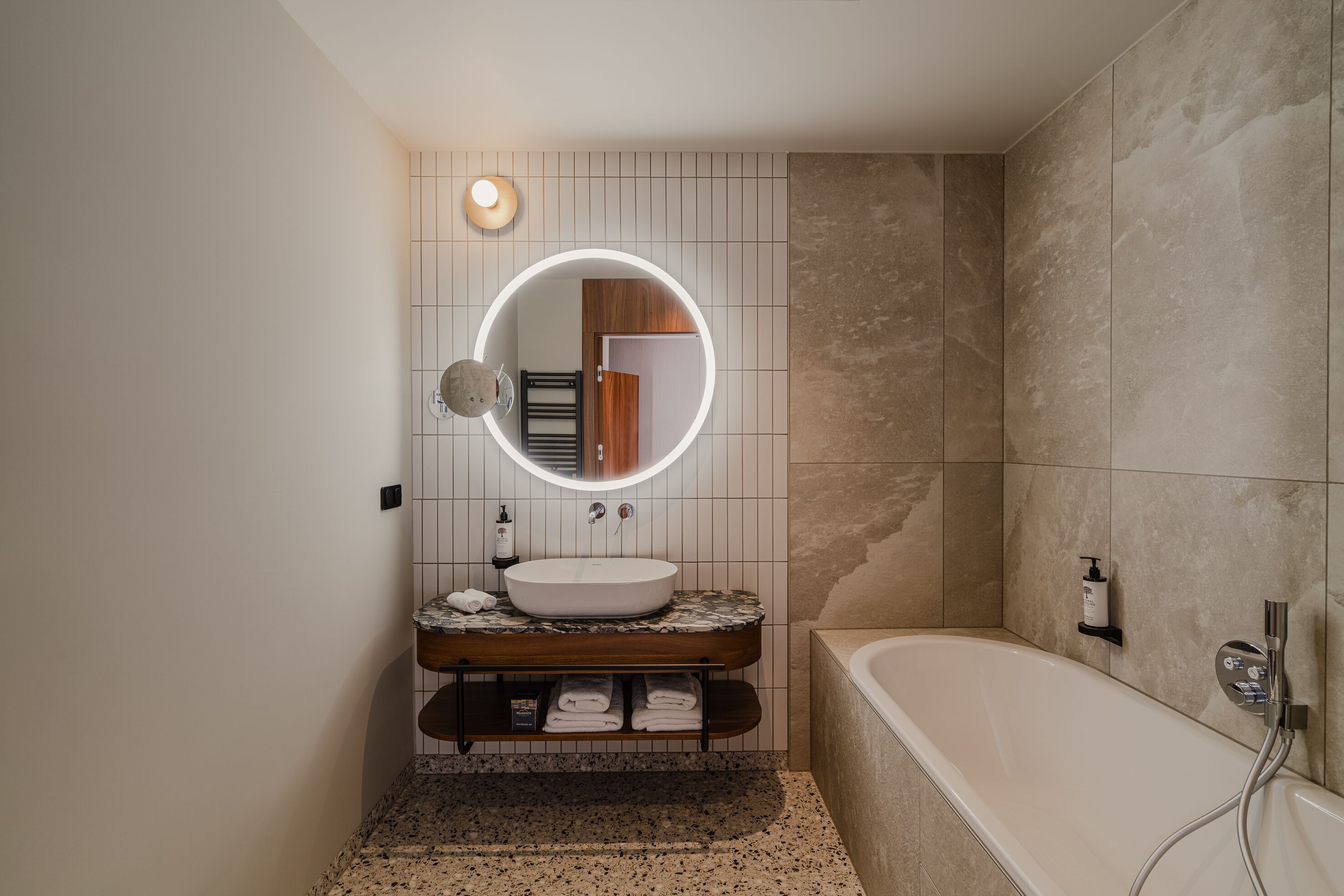 A modern bathroom with a freestanding bathtub, a round mirror with a light, and a marble countertop.