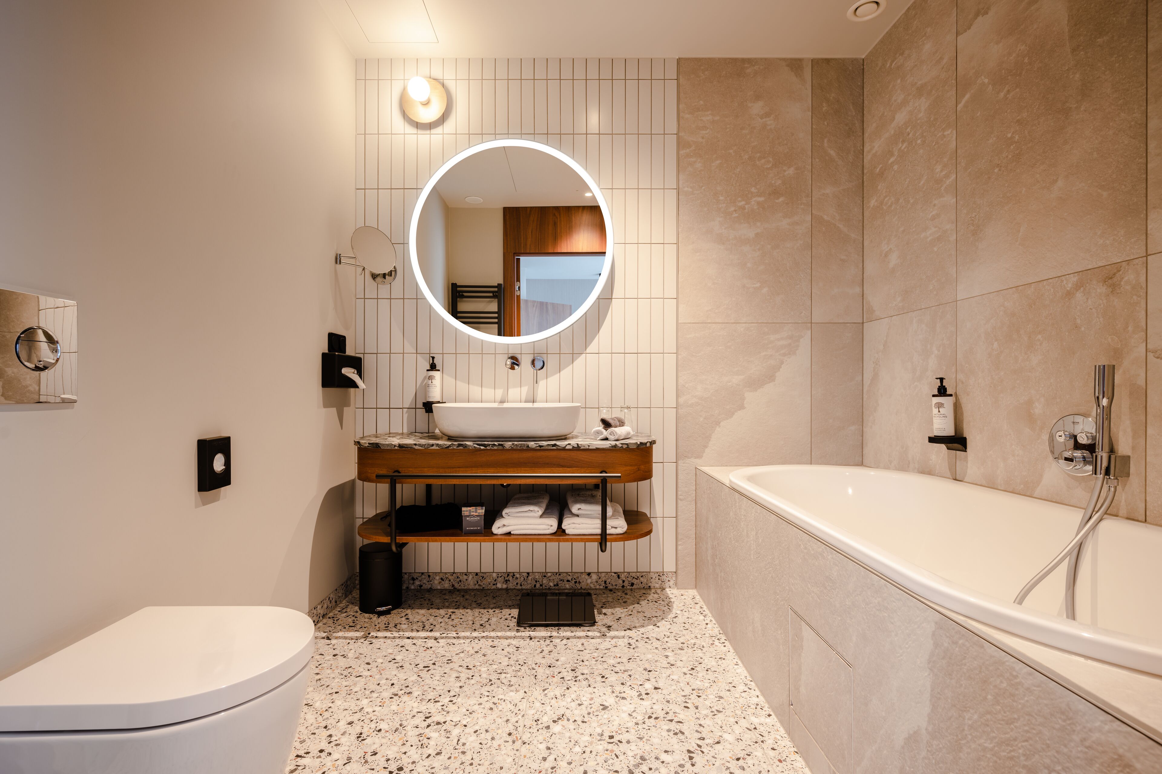 A modern bathroom with a freestanding bathtub, a round mirror, a sink with a wooden countertop, and a toilet.