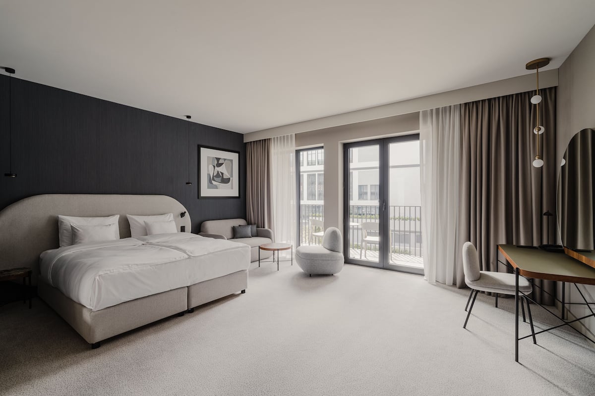 Une chambre d’hôtel moderne, dotée d’un grand lit, d’un canapé, d’un bureau et d’une chaise. Une grande fenêtre, agrémentée de rideaux, offre une vue sur l’extérieur.