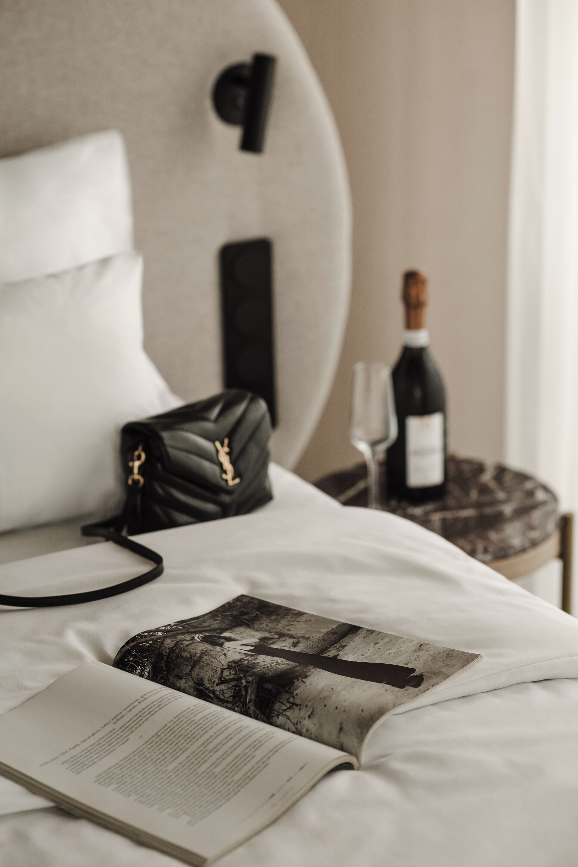 A neatly made bed with a book, a black handbag, and a bottle of wine on a bedside table.