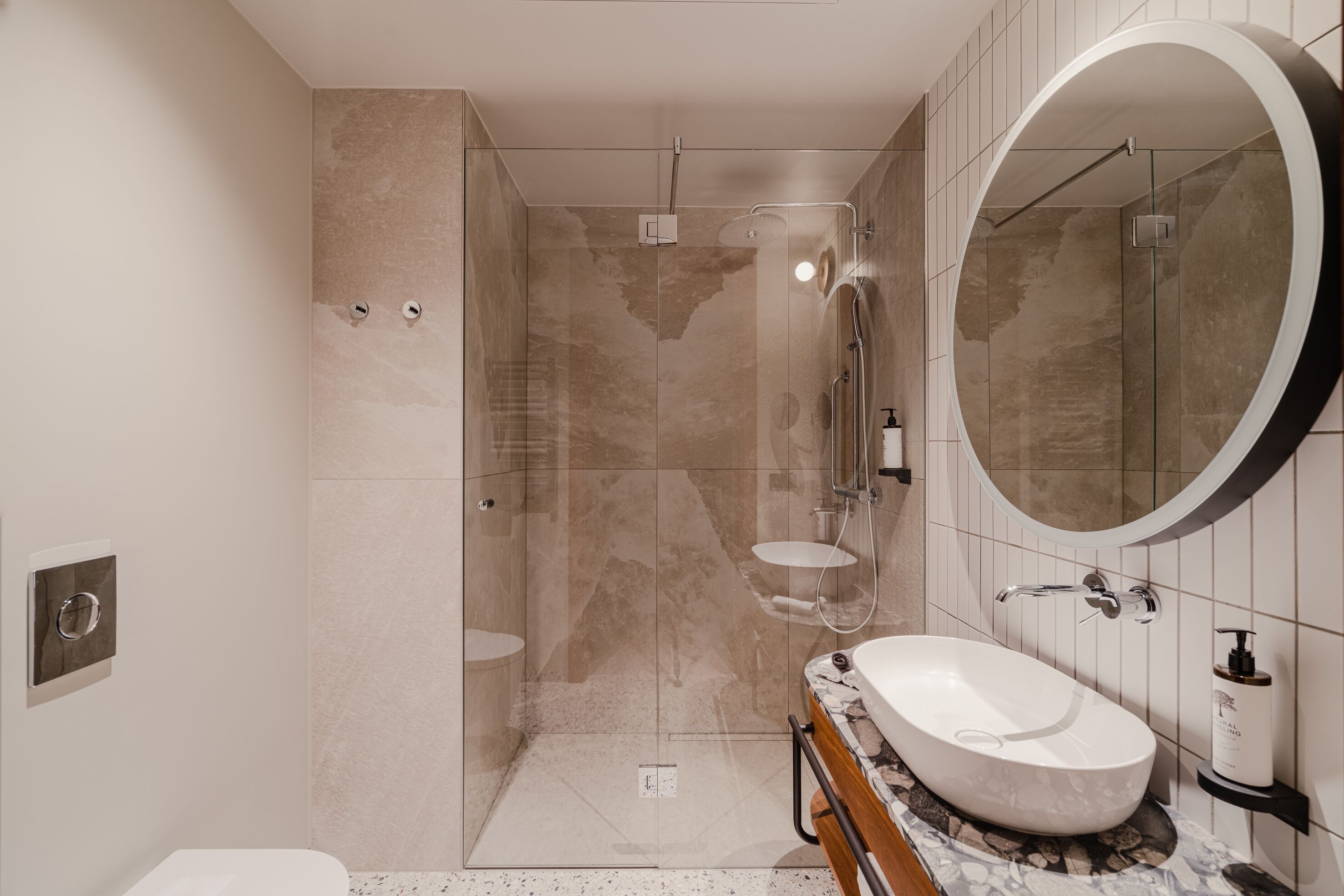 A modern bathroom with a glass-enclosed shower, a large oval mirror, and a marble sink.