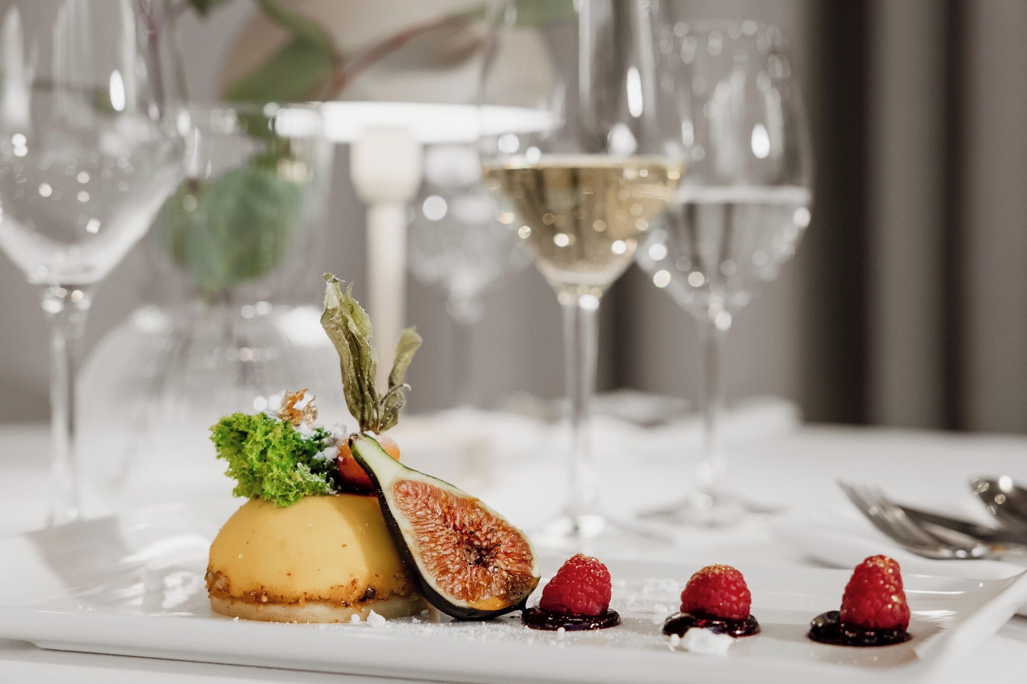 Une assiette de dessert avec une figue, des framboises et une petite décoration verte, accompagnée d'un verre de vin blanc.