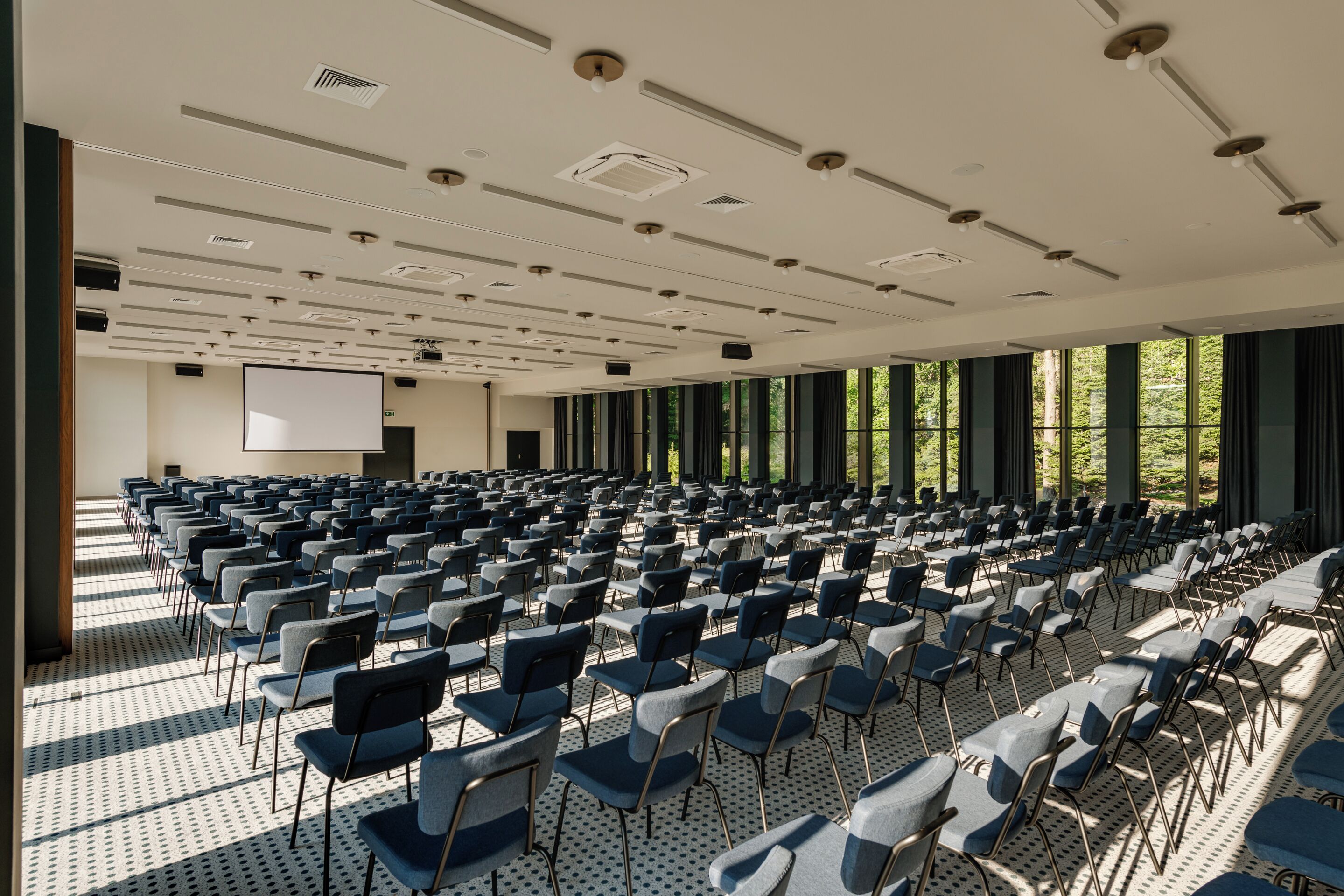Een congreszaal met rijen stoelen, een projectiescherm en grote ramen met gordijnen.