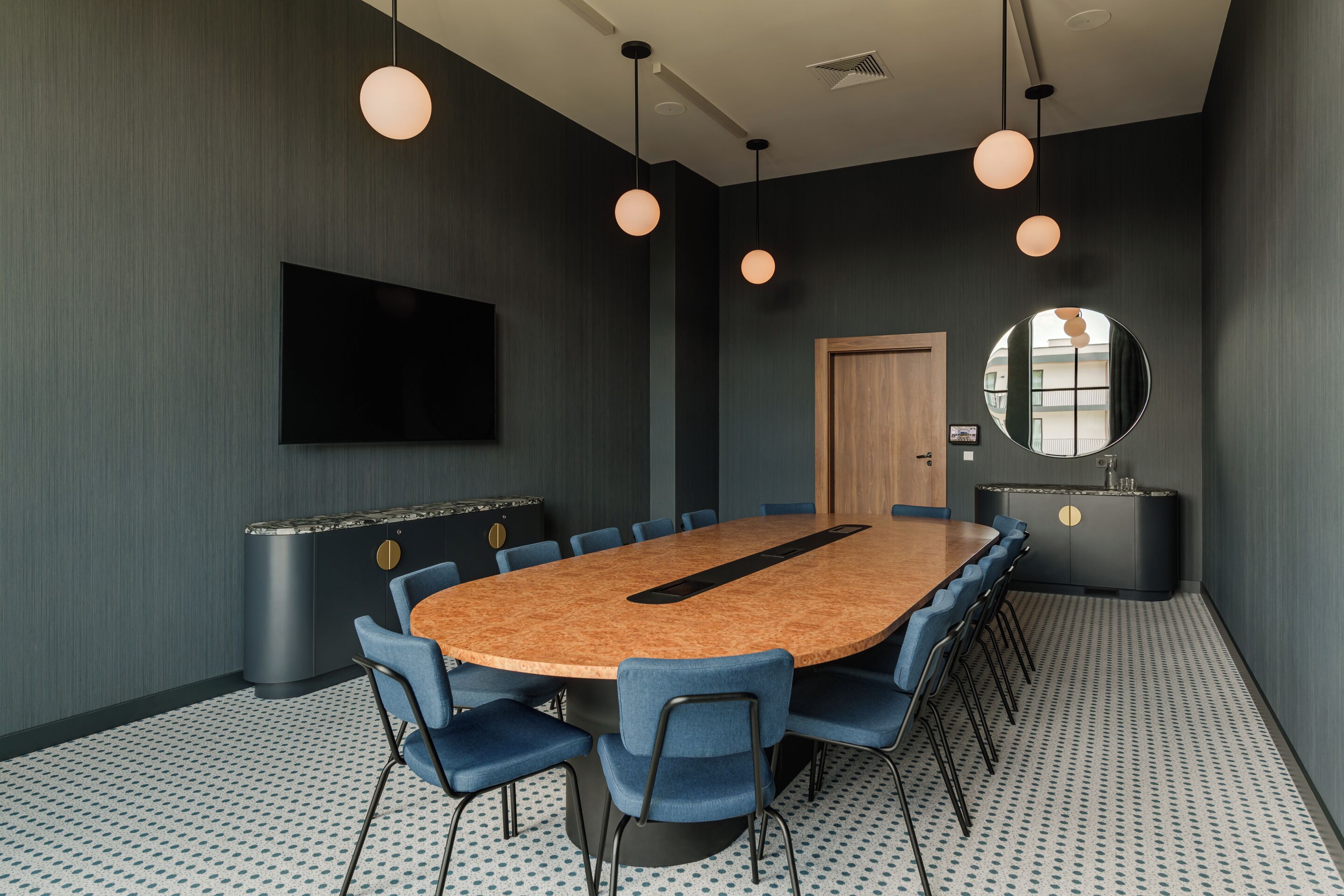 Une salle de conférence équipée d’une table ronde, de chaises bleues, d’un téléviseur à écran plat et de luminaires suspendus.