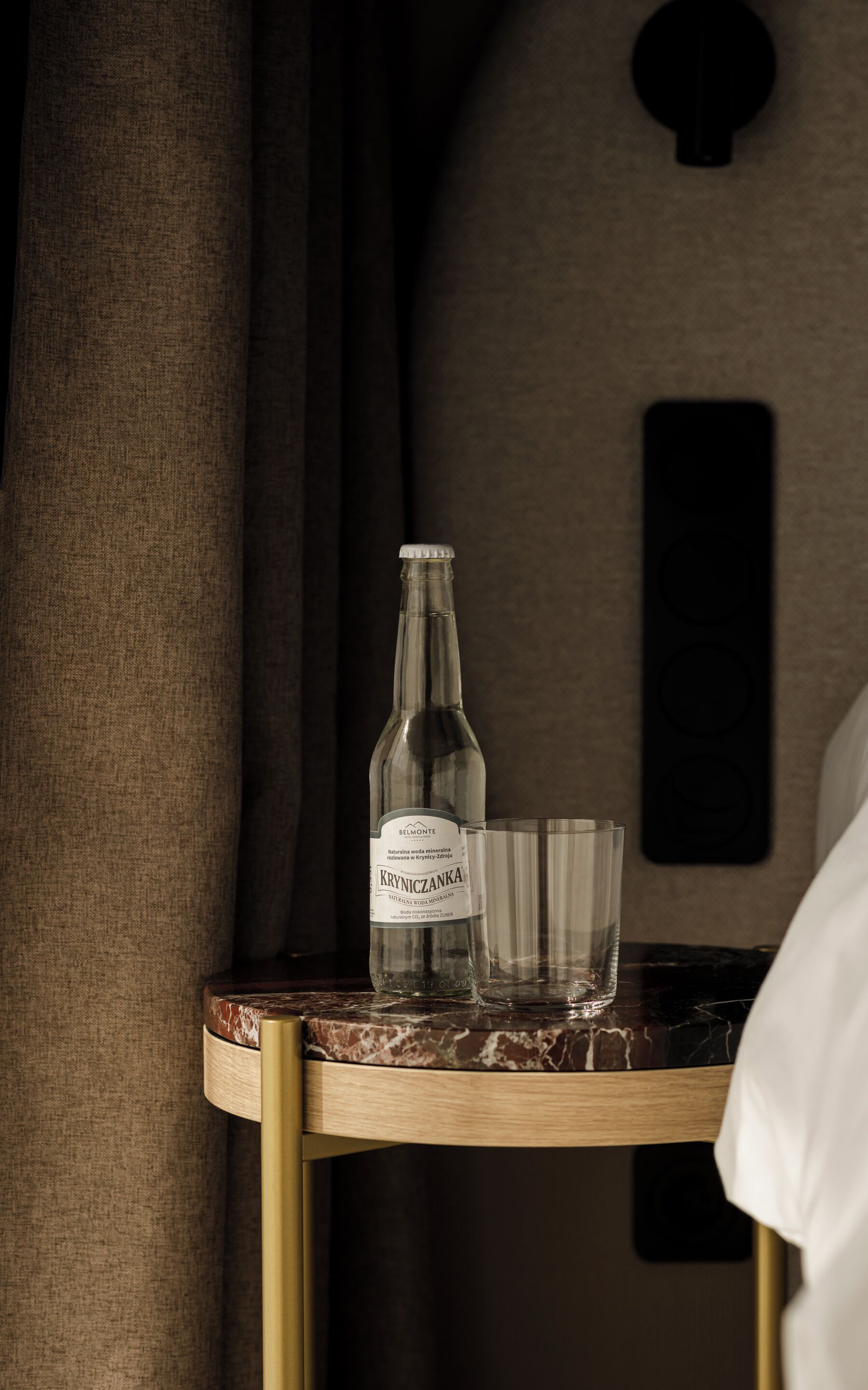 Una botella de agua mineral Belmont Kryniczanka y un vaso sobre una mesa de mármol en una habitación con pared texturizada y un ojo de cerradura.