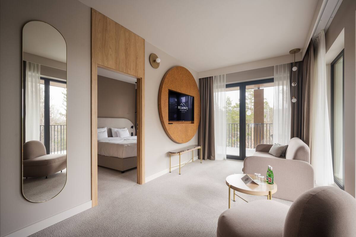Une chambre d’hôtel moderne équipée d’un lit, d’un canapé, d’une petite table avec un ordinateur portable et des boissons, d’un téléviseur fixé sur un panneau en bois et d’un grand miroir.