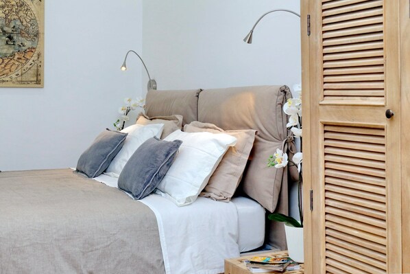 A neatly made bed with a beige and white duvet, a wooden door with horizontal slats, and a wall-mounted lamp.