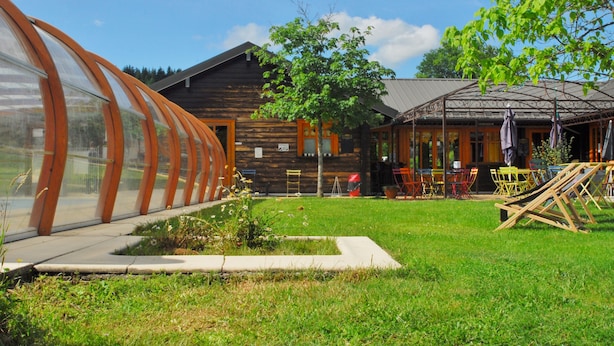 A wooden cabin with a glass greenhouse structure, outdoor seating area, and a small pond.