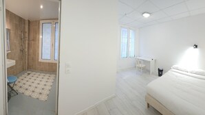 A bedroom with a bed, a desk, and a chair. There is a shower area with a tiled floor and a small stool. - Hotel CORONA Lourdes (Lourdes)
