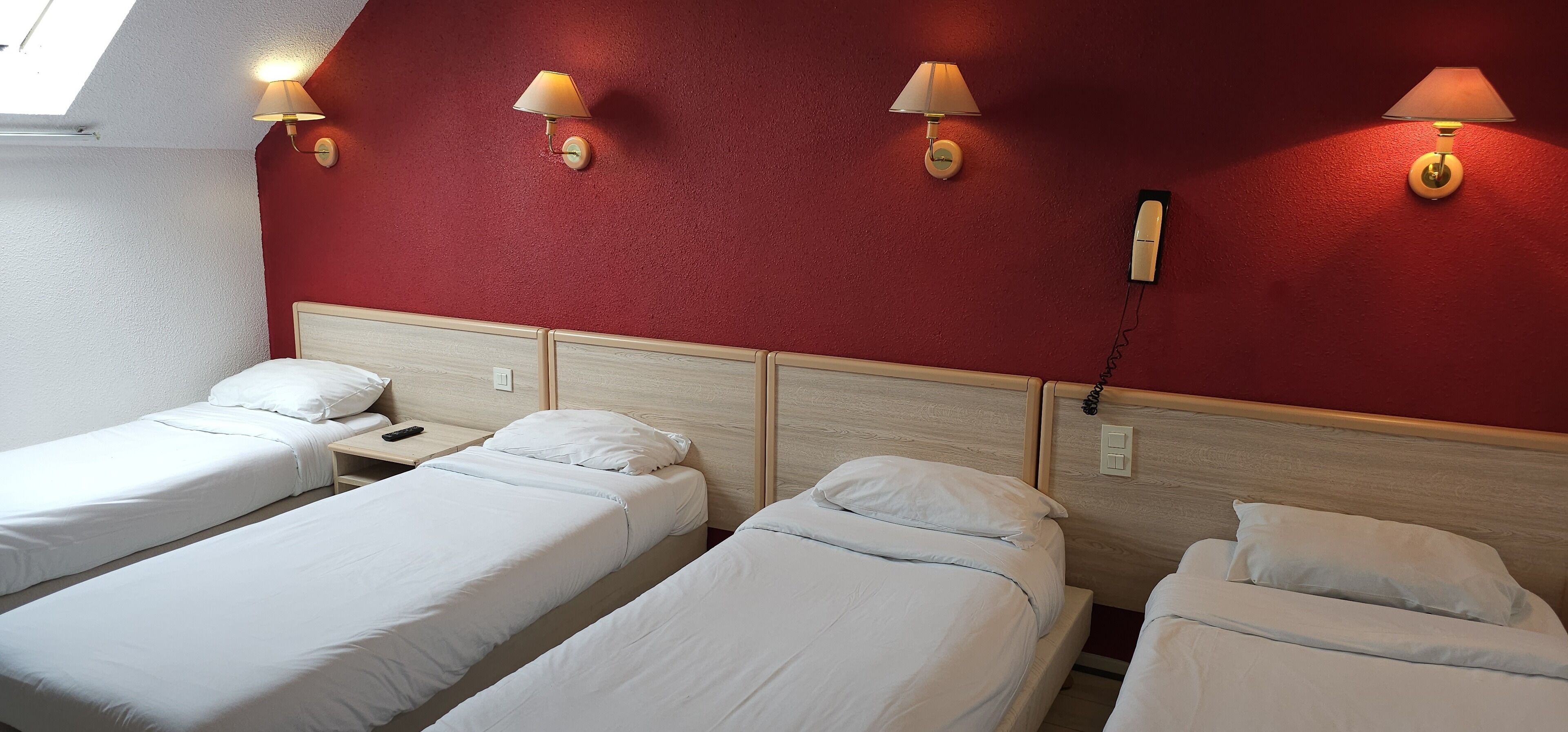 A hotel room with three single beds, each with white bedding and a wooden headboard. There are wall-mounted lamps with beige shades on the red-painted wall.