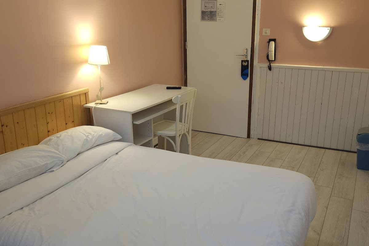 Ett hotellrum med en säng, ett skrivbord, en stol, en lampa, en telefon och en papperskorg.