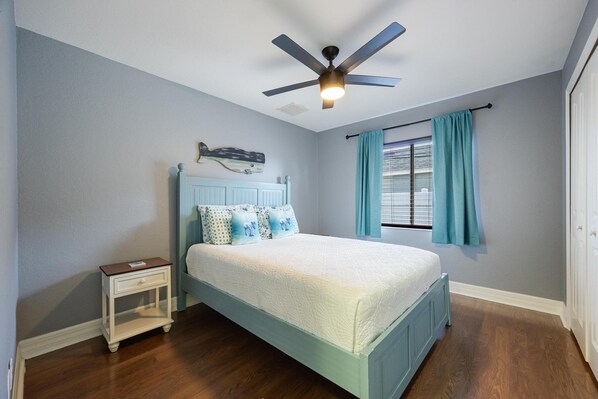 A bedroom with a bed, a nightstand, a ceiling fan, a window with curtains, and a wall-mounted decoration.