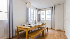 A dining area with a wooden table set for four, two pendant lights, and a view of the city through large windows. - Appartement8P/Full Equipped/Bonnenouvelle/Bourse (Paris)