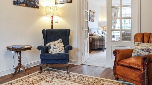 A room with a blue armchair, a wooden side table, a patterned rug, and a door leading to another room with a bed and a window. - 3 bedroom accommodation in Keswick (Keswick)