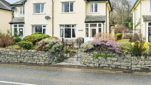 A two-story house with a stone wall and a gate, surrounded by flowering plants and a clear sky. - 3 bedroom accommodation in Keswick (Keswick)