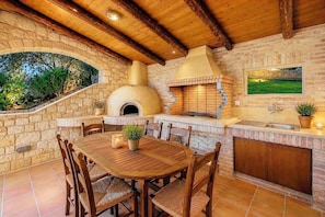 Cucina esterna con pareti in pietra, dotata di forno a mattoni, tavolo e sedie.
