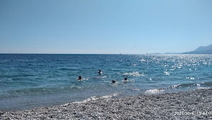 Una spiaggia con ciottoli, acque cristalline e montagne in lontananza.