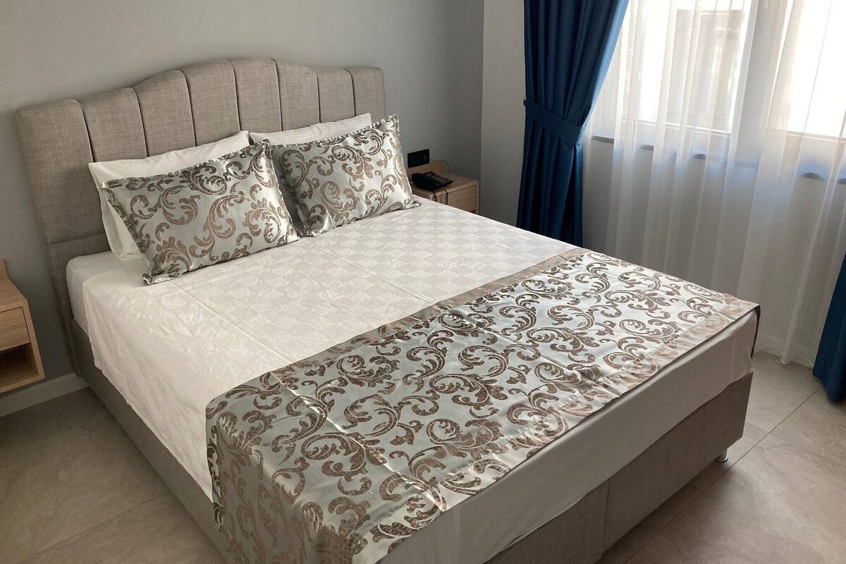 Una cama bien hecha con una colcha y almohadas estampadas, una mesita de noche con una lámpara y una ventana con cortinas.